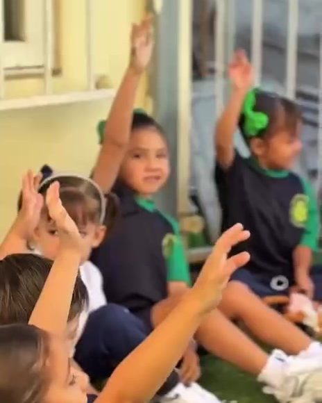 Niñas y niños de preescolar sentados en el pasto levantando la mano para contestar
