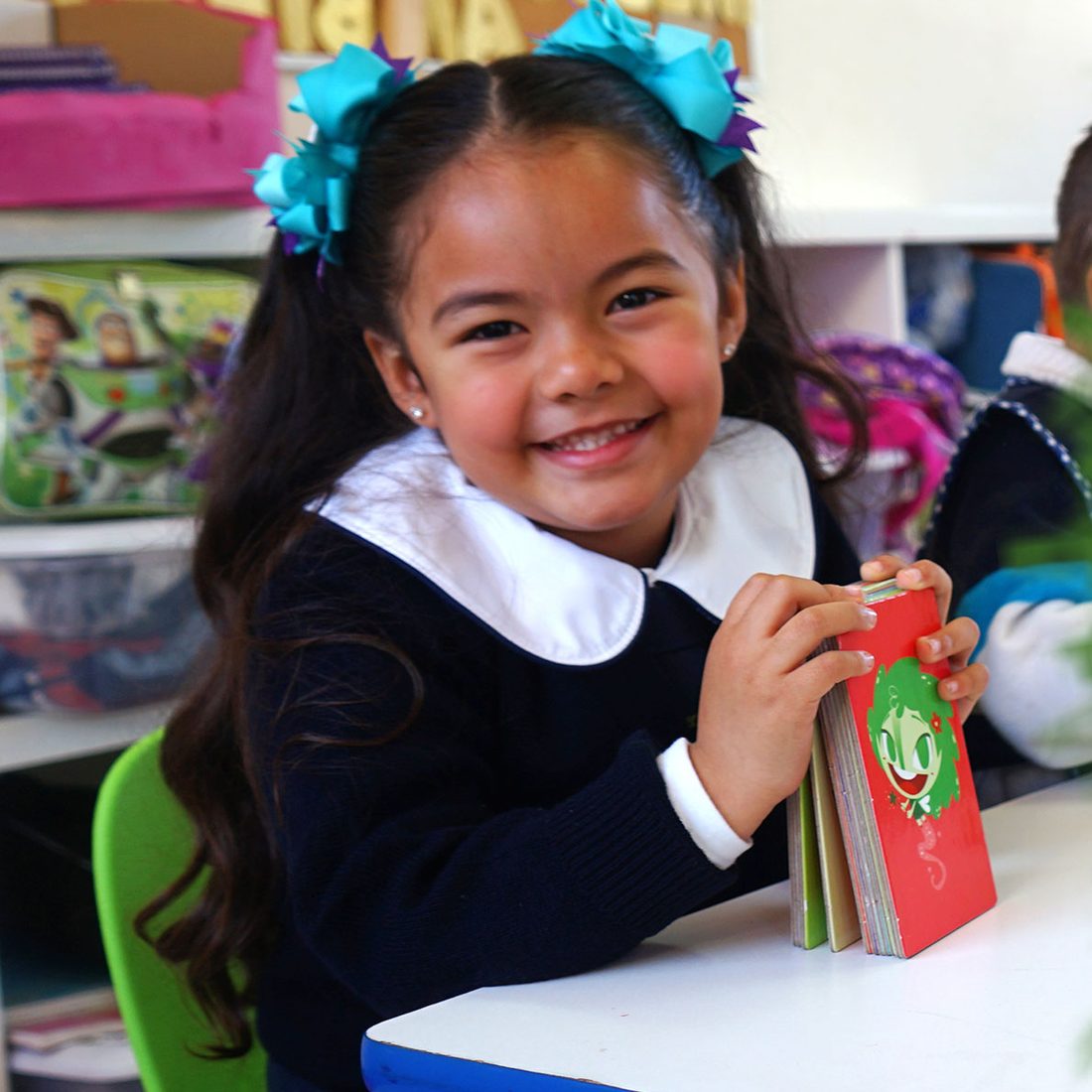Alumna de preescolar de Colegio Encinos con uniforme azul marino y moños turquesa, sonriendo con un libro de cuentos en las manos