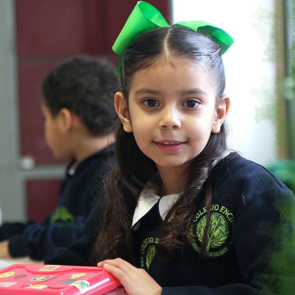 Niña de preescolar con moño verde y uniforme de Colegio Encinos sentada en su mesa de trabajo del salón