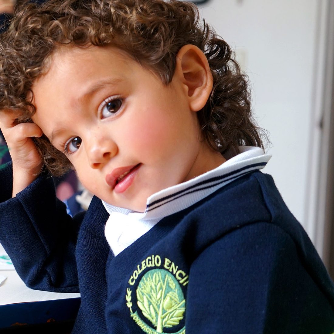 Niño de maternal de rizos con el suéter del uniforme de Colegio Encinos, recargando la cabeza en la mano y mirando a la cámara