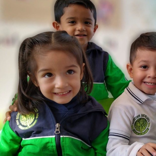 Tres alumnos de maternal y preescolar de Colegio Encinos, con chamarra verde y playera blanca del uniforme, sonriendo en el salón