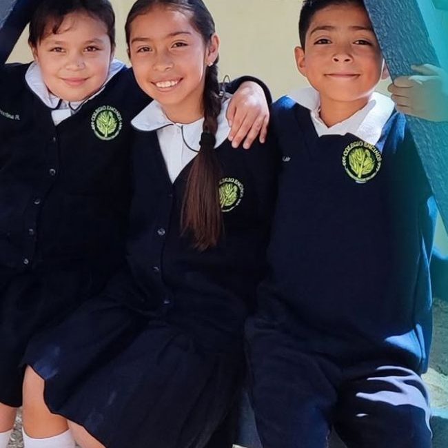 Tres alumnos de primaria de Colegio Encinos con uniforme azul marino abrazados en los juegos del patio, en Tecate